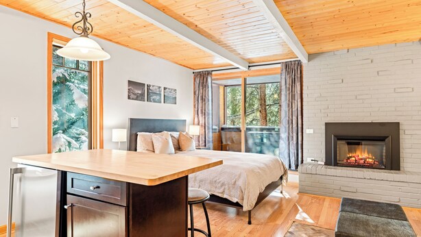 A modern bedroom with a wooden ceiling, a fireplace, a bed, and a kitchen area with a refrigerator and bar stools. - Studio in Great Area, Easy Walk to Town & Peak 9 (Breckenridge)