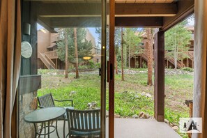 Blick von einem Zimmer mit einer Glastür, die auf eine Außenpatio führt. Die Patio hat einen Tisch und Stühle, und der Ausblick umfasst ein mehrgeschossiges Gebäude mit einer Holzterrasse.