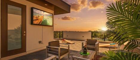A modern outdoor seating area with a television screen, a glass door, and a view of the sunset.