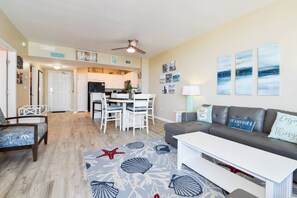 Un vaste salon avec un canapé gris, une table basse blanche et un tapis orné de motifs d’étoiles de mer. On aperçoit à l’arrière-plan un coin repas avec des chaises blanches et une table.