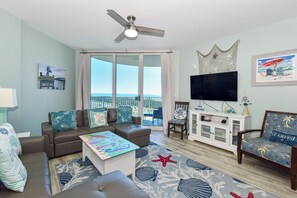 A living room with a sofa, coffee table, TV, and a view of the ocean through large windows.