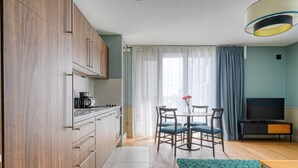 A modern kitchen with wooden cabinets, a dining area with blue chairs, a TV, and a large window with curtains.