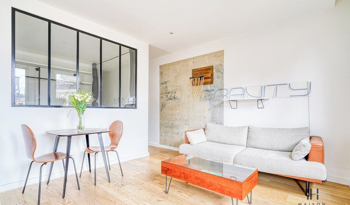 A modern living room with a glass coffee table, a grey sofa, and a dining area with two chairs.