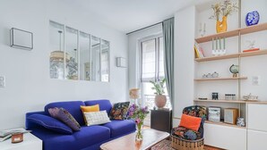 Una sala moderna con un sofá azul, un sillón estampado, una estantería y un amplio ventanal con cortinas.