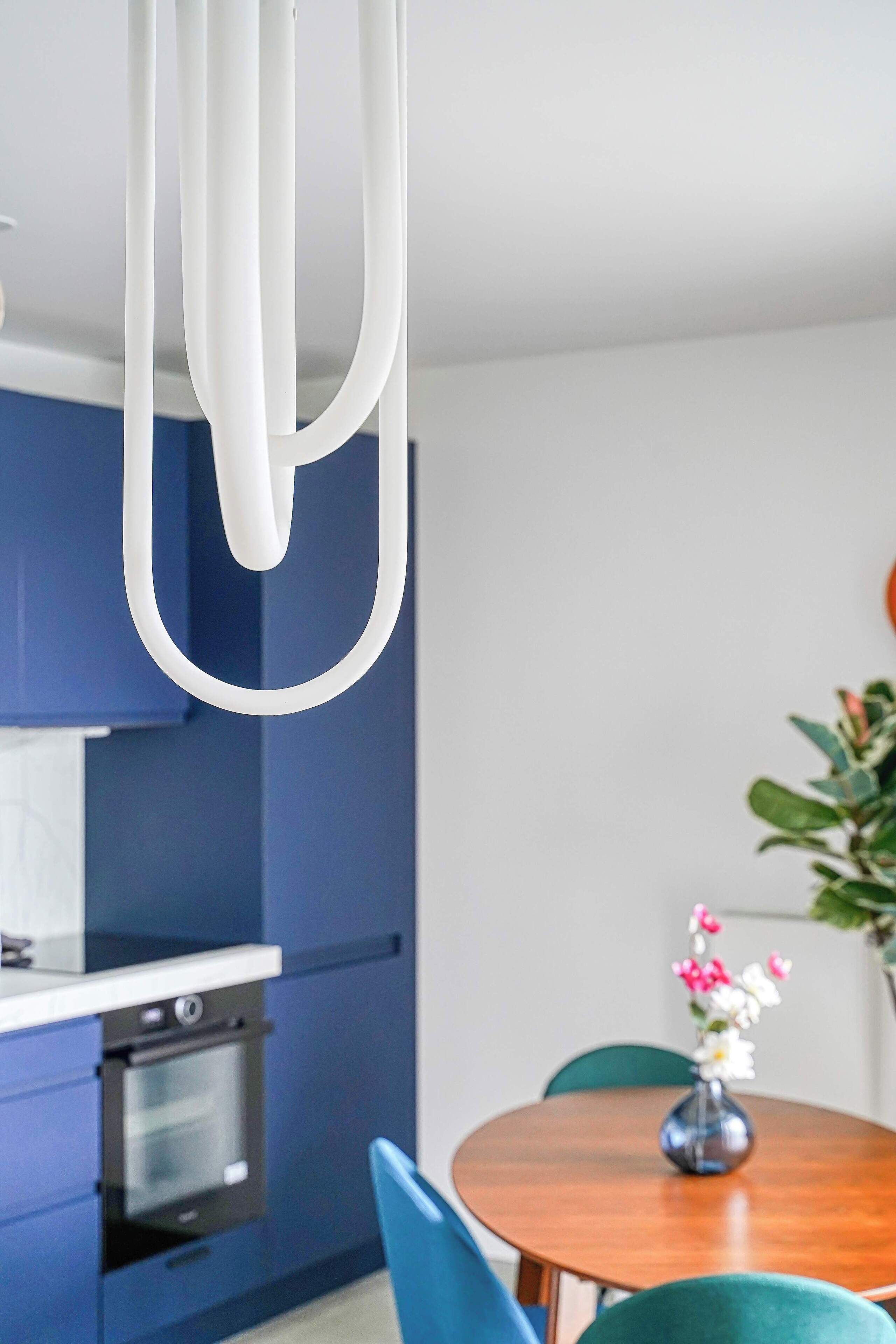 Une cuisine moderne avec des meubles bleus, un luminaire suspendu blanc et un coin repas comprenant une table en bois et des chaises vertes.