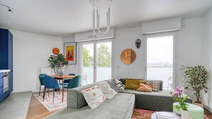 Una sala de estar moderna con un sofá gris, una mesa de comedor con sillas, una zona de cocina y ventanas amplias.