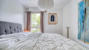 Un dormitorio con cabecera capitonada, cama grande con cubrecama acolchado, ventana con cortinas, una obra de arte colgada en la pared y una lámpara de pie.