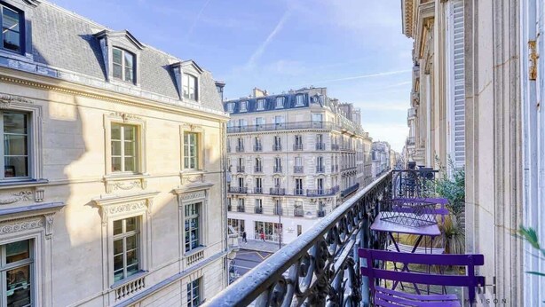 A narrow street with buildings on either side, a balcony with a railing, and a purple bench. - Invalides - The Collector (Paris)