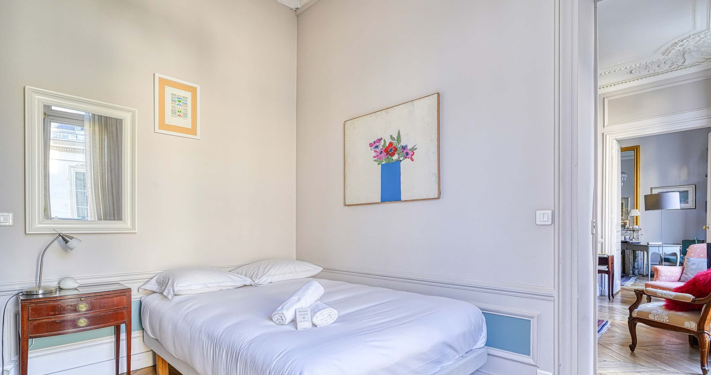 A bedroom with a bed, a nightstand, a framed picture, and a door leading to another room.