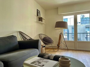 Een moderne woonkamer met een grijze bank, een rieten fauteuil, een vloerlamp en uitzicht door een groot raam.