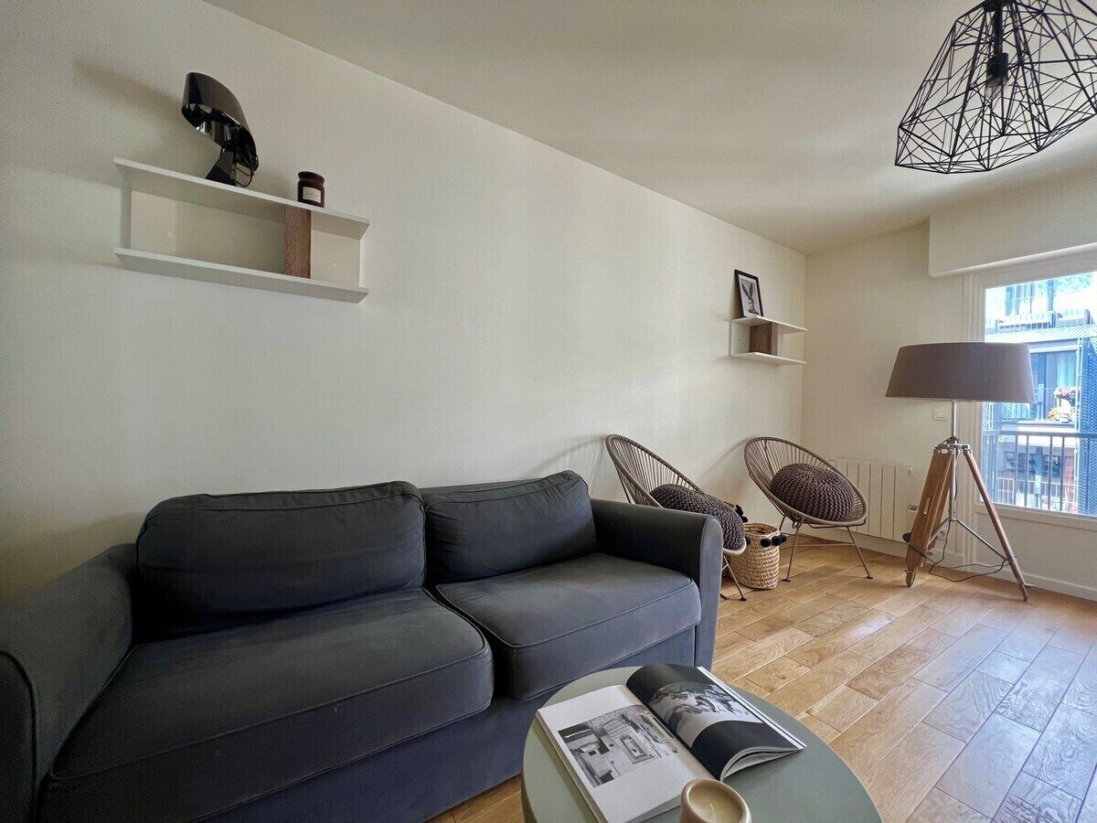 Een moderne woonkamer met een donkergrijze bank, een rond salontafeltje met tijdschriften, een rieten fauteuil, een vloerlamp en een boekenkast met decoratieve objecten.