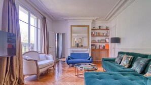 Un salón con un sofá blanco, un sillón azul y un sofá verde azulado. Hay una chimenea con un espejo encima y una estantería con libros y objetos decorativos.