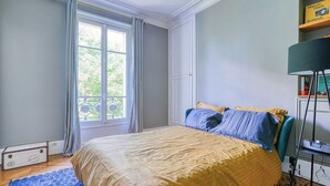 Un dormitorio con cama, ventana con cortinas, estantería y lámpara de pie.