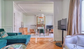 A spacious living room with a teal sofa, a white armchair, and a coffee table. There is a fireplace with a mirror above it, and a dining area with chairs and a table. A television is mounted on the wall, and a large window with curtains is visible.