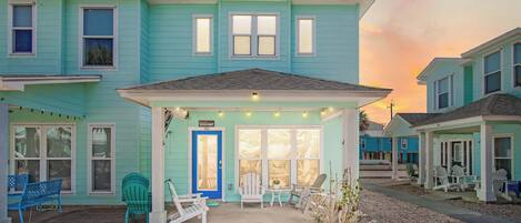 A turquoise two-story building with a covered porch, outdoor seating, and a blue door.