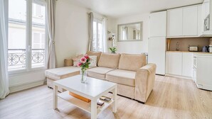 A living room with a beige sofa, a white coffee table with a vase of flowers, and a kitchen area with white cabinets and appliances. - Ternes district - The Dreamer (Paris)