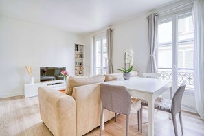 Una sala moderna con un sofá color beige, una mesa de comedor y un televisor. Se observa una estantería y una planta en maceta.