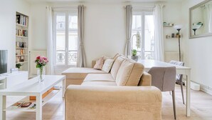 A modern living room with a beige sofa, a white coffee table, and a dining area with a white table and chairs. There is a bookshelf filled with books and a vase with flowers on the coffee table. - Ternes district - The Dreamer (Paris)