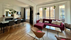 Ein geräumiges Wohnzimmer mit einem roten Sofa, einem Essbereich mit Tisch und Stühlen sowie großen Fenstern mit Stadtblick.