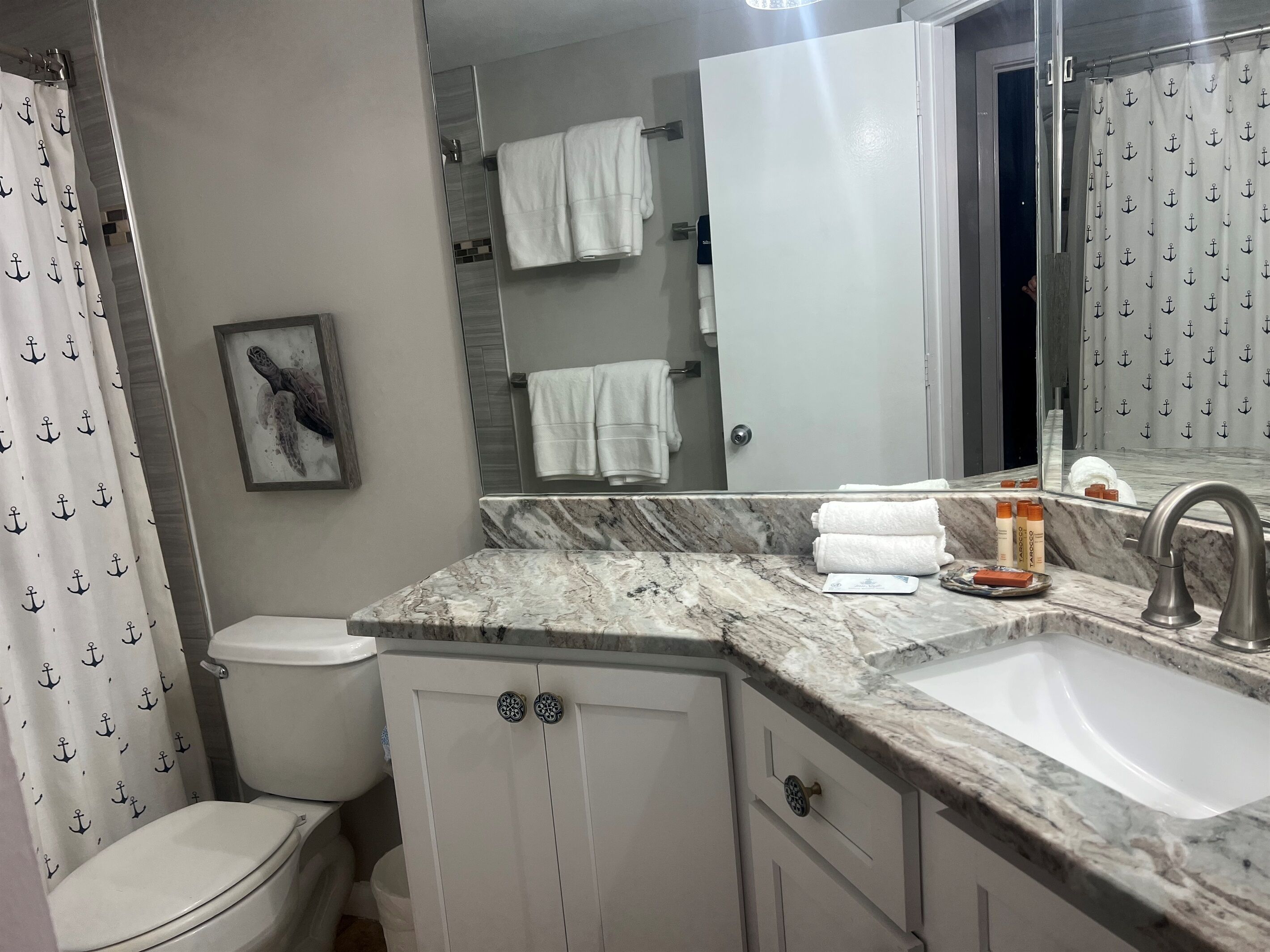 A bathroom with a marble sink, white cabinets, a toilet, and a shower curtain with anchor patterns.