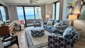 A living room with a sofa, ottoman, and chair, a TV, a ceiling fan, and a view of the ocean through large windows.
