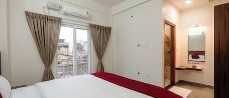 Habitación de hotel con cama, ventana con cortinas, unidad de aire acondicionado, espejo y puerta que da a un baño.