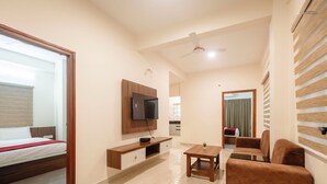 A modern hotel room with a bed, a TV mounted on a wooden cabinet, a small table, a sofa, and a ceiling fan. - Aetheria Service Apartments (Chikkamagaluru)