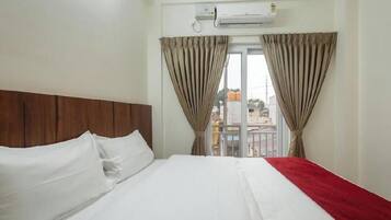 A hotel room with a bed, curtains, an air conditioning unit, and a view of the outside.