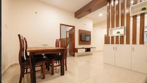 A dining area with a wooden table and chairs, a TV mounted on the wall, and a cabinet with shelves. - Aetheria Service Apartments (Chikkamagaluru)