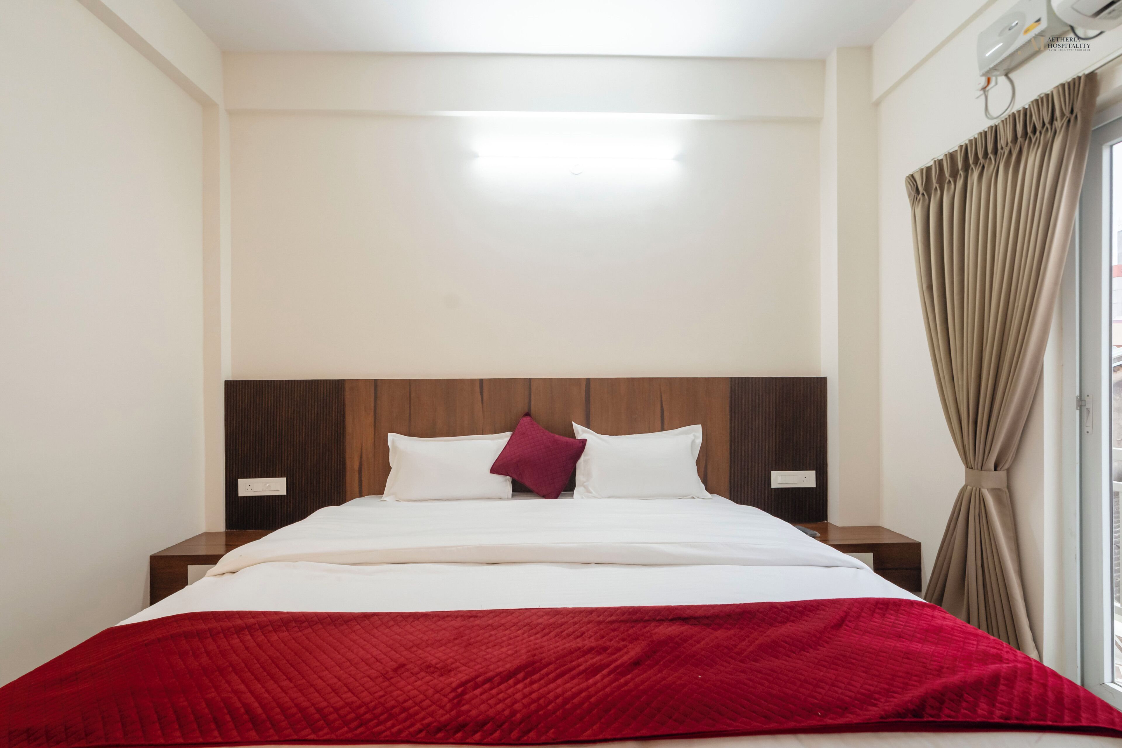 A hotel room with a bed, a red blanket, wooden headboard, two bedside tables, and a window with curtains.
