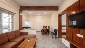 Una sala de estar moderna con un sofá, una mesa de centro de madera, un comedor con sillas, un televisor montado en la pared y una cocina al fondo.