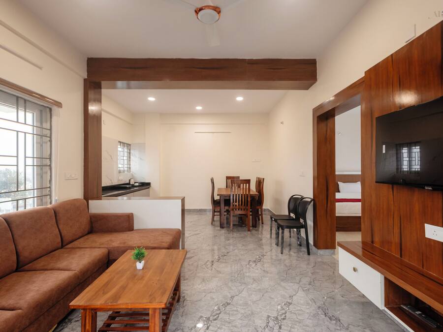 Una sala de estar moderna con un sofá, una mesa de centro de madera, un comedor con sillas, un televisor montado en la pared y una cocina al fondo.