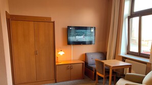 Habitación de hotel con armario de madera, una mesita, una silla, televisor montado en la pared y una ventana con cortinas.