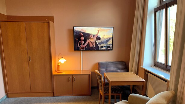 Habitación pequeña con un armario de madera, una mesa con una silla, televisor montado en la pared y una ventana con cortinas.