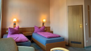 Habitación de hotel con dos camas, cada una con cabecero de madera y colchón azul. Hay dos mesitas de noche con lámparas, y una puerta de madera con manija.