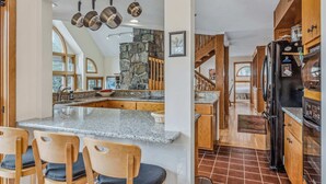 A kitchen with granite countertops, a stone accent wall, hanging pots, and a modern refrigerator. - Whitman Brook Overlook House with Game Room (Quechee)