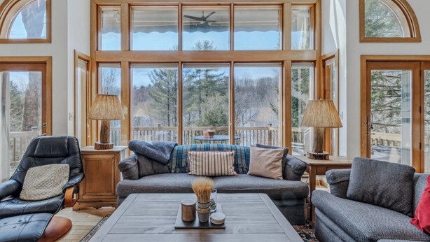 A spacious room with a large window, a sofa, a chair, a table, and a lamp. - Whitman Brook Overlook House with Game Room (Quechee)