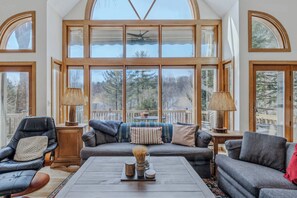 A spacious room with a large window, a sofa, a chair, a table, and a lamp. - Game Room - Whitman Brook Overlook (Quechee)