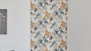 Un coin repas avec une table en verre et quatre chaises, un canapé gris et un mur orné d’un motif de feuilles tropicales.