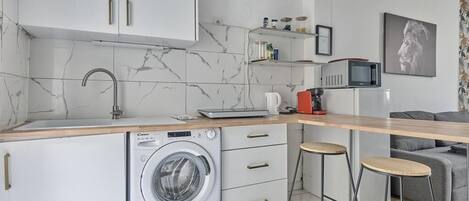 A compact kitchen with a washing machine, a microwave, and a coffee maker. There are two stools and a grey sofa in the living area.