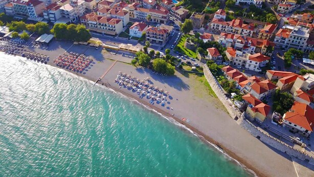 An aerial view of a coastal town with a beach lined with umbrellas and sun loungers, surrounded by residential buildings with red roofs. - Nafpaktos Cozy Nest, Homely Vibes near the Beach (Paleopanagia)