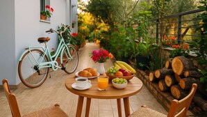 A breakfast setup with croissants, coffee, juice, and fruit on a patio with a bicycle and plants in the background. - Nafpaktos Cozy Nest, Homely Vibes near the Beach (Paleopanagia)