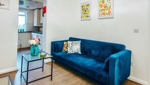 Una sala de estar moderna con un sofá azul, una mesa de centro de vidrio y una lámpara de techo circular única. Hay dos cuadros en la pared y un florero con flores sobre una mesita auxiliar.