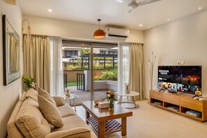 Una sala de estar moderna con sofá, mesa de centro y televisor. Tiene un balcón con plantas y vista a zonas verdes.