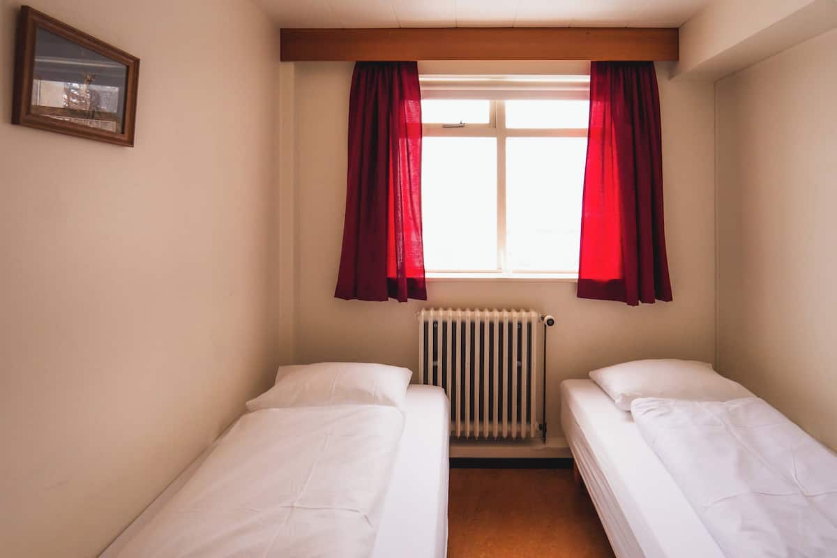 A small, clean room with two single beds, a window with red curtains, a radiator, and a framed picture on the wall.