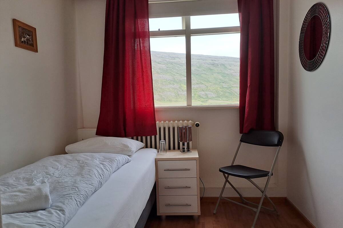 A bedroom with a bed, a chair, a radiator, and a window with curtains.