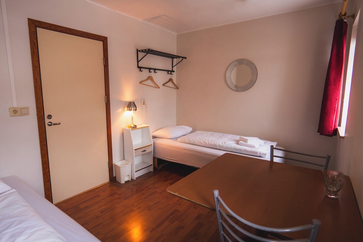 A small, tidy room with a wooden floor, a single bed, a table with a chair, a bedside table with a lamp, and a wall-mounted mirror.