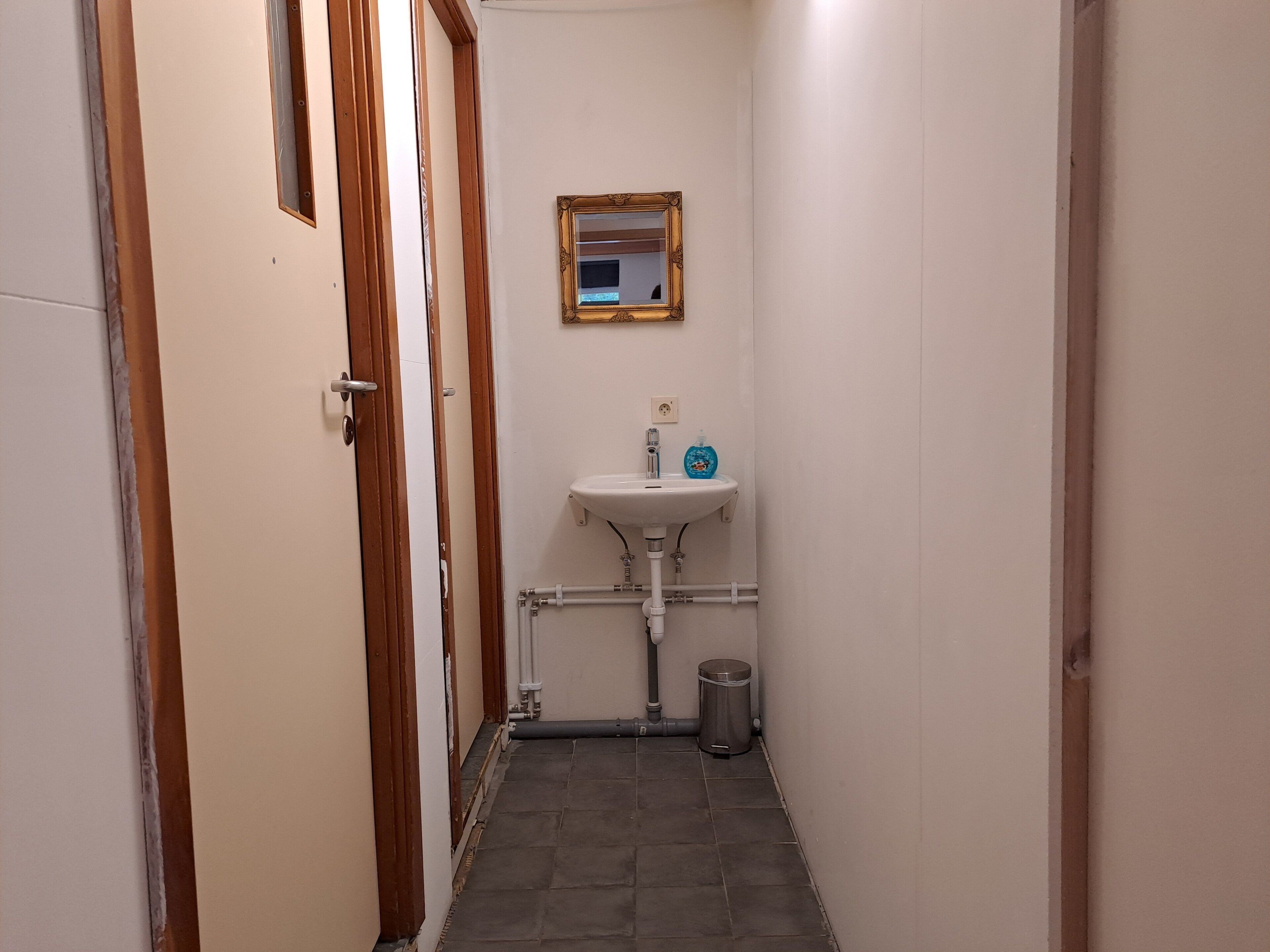 Un couloir étroit mène à une salle de bain équipée d’un lavabo blanc, d’un miroir et d’une poubelle.