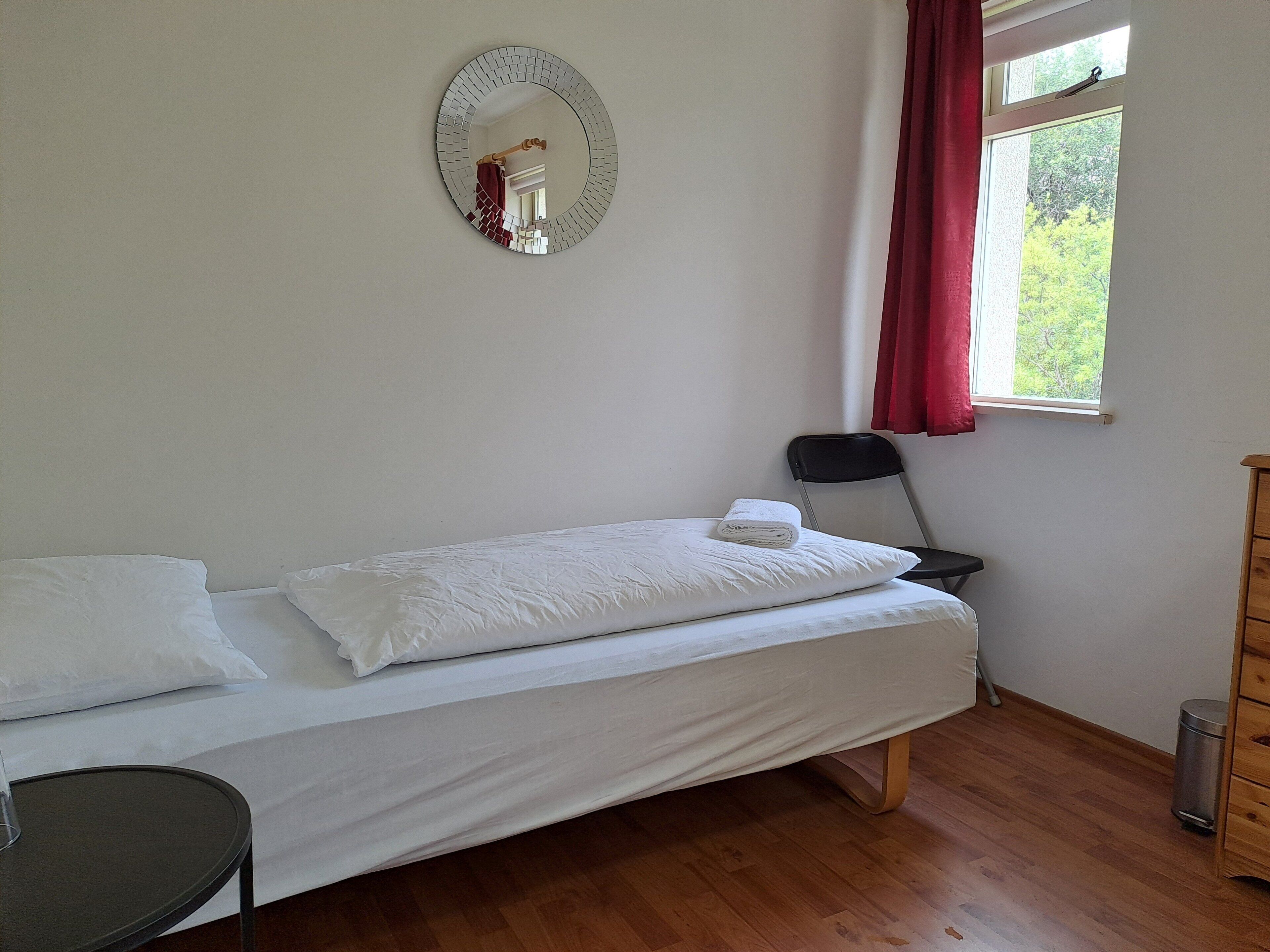 Un lit simple avec du linge de lit blanc, un miroir rond accroché au mur, une chaise, une petite table, une fenêtre avec des rideaux rouges et une commode en bois.