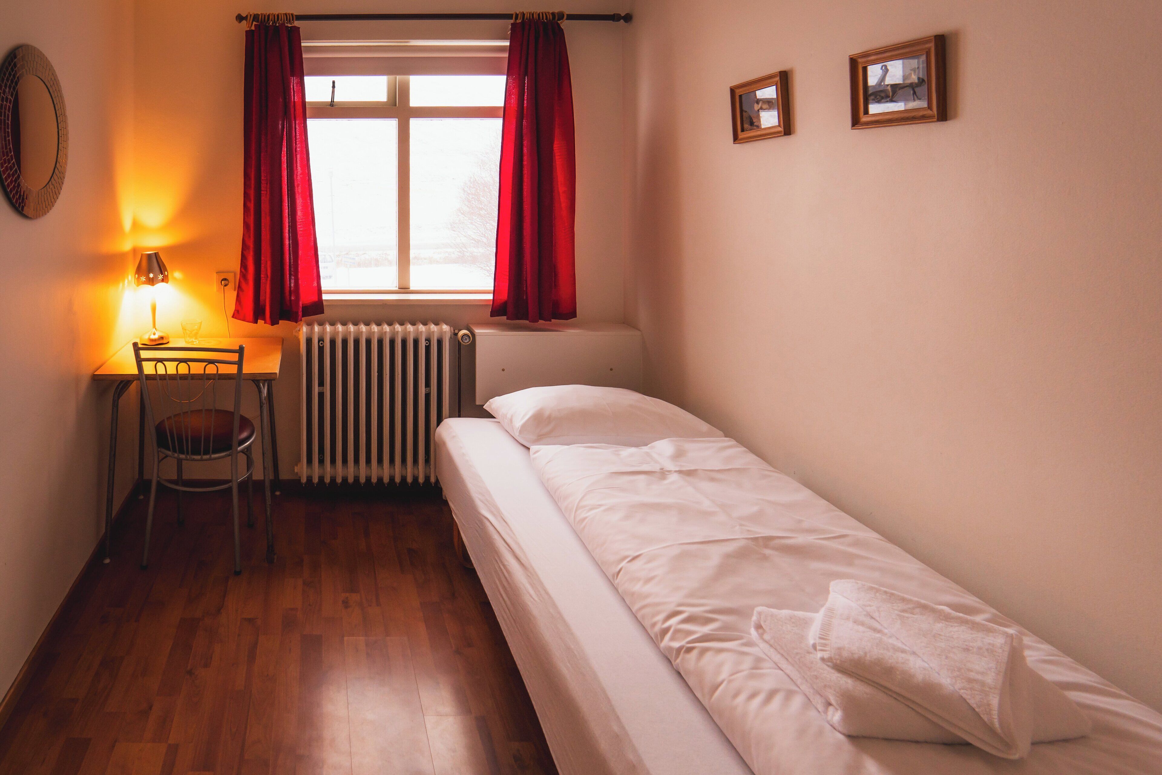 A small, single-bed room with a wooden floor, a desk with a chair, a radiator, and a window with red curtains.